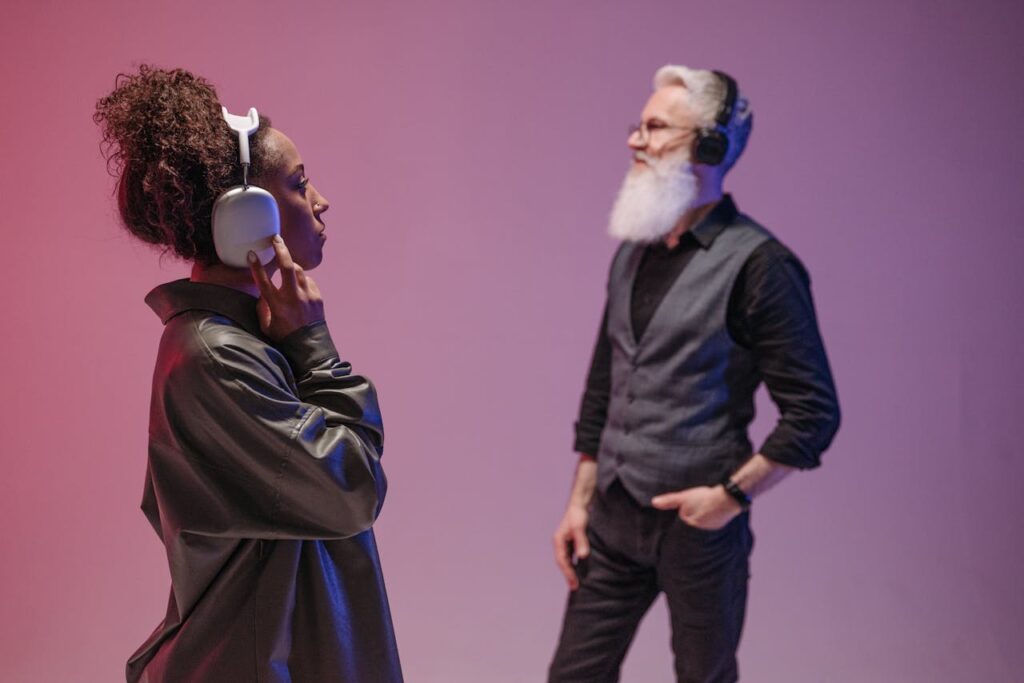 A black woman and a bearded senior man enjoy music with headphones in a modern studio setting.