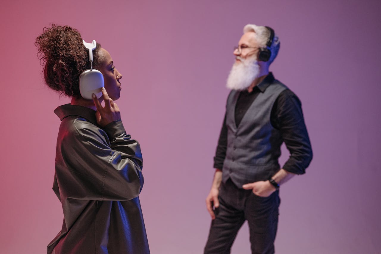 A black woman and a bearded senior man enjoy music with headphones in a modern studio setting.